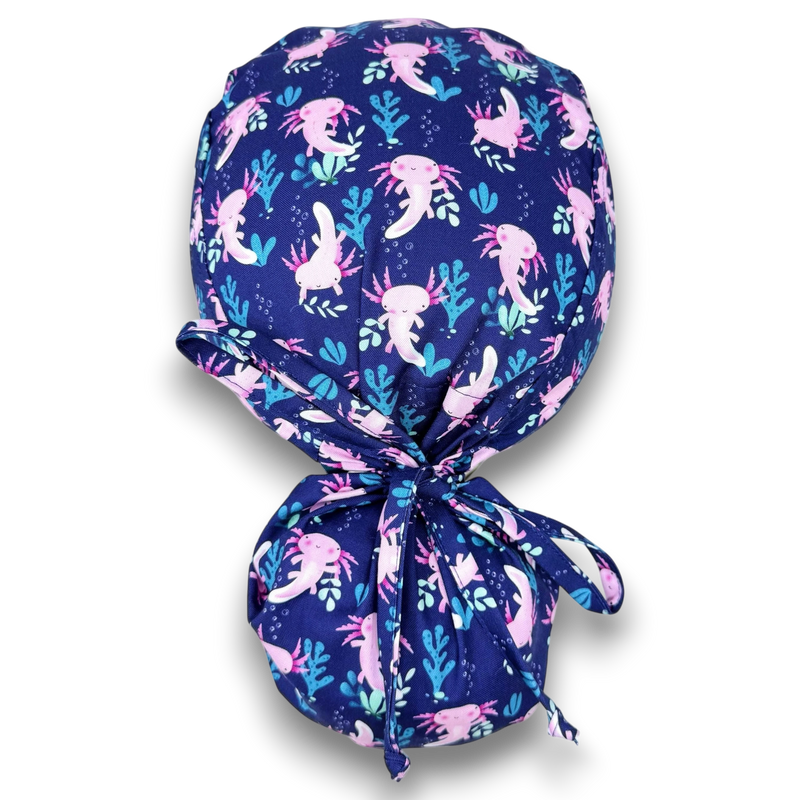 Paradise Caps navy blue ponytail scrub cap with pink axolotl and blue plant print. 100% cotton.